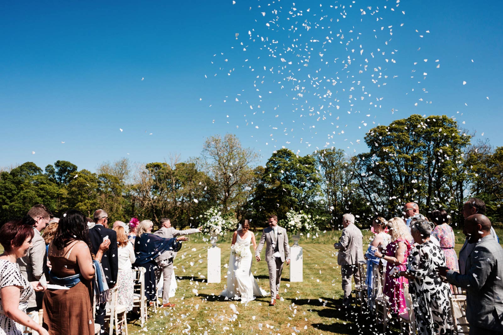 The image shows a bride and groom walking down the aisle in the grounds of Yorebridge House in Bainbridge. Their exclusive use house wedding gave them freedom to use the grounds, house and allow their day to flow naturally. With confetti falling from the clear blue skies, the Yorkshire Dales offered the perfect location for their wedding.