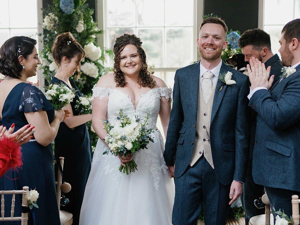 The image shows a bride and groom walking down the aisle during their intimate winter wedding ceremony at Yorebridge House. The bride is wearing a drop-shoulder dress with floral appliqués and deep-plunging neckline. The groom is wearing a dark blue suit with a beige vest and tie.
