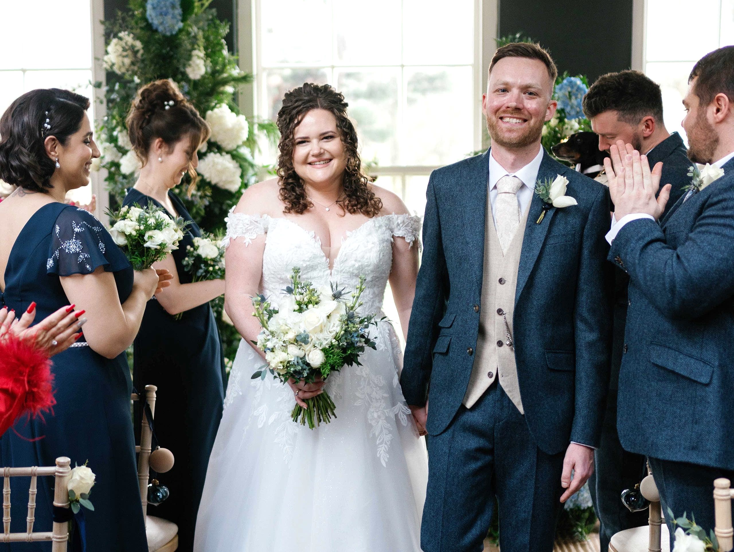 The image shows a bride and groom walking down the aisle during their intimate winter wedding ceremony at Yorebridge House. The bride is wearing a drop-shoulder dress with floral appliqués and deep-plunging neckline. The groom is wearing a dark blue suit with a beige vest and tie.