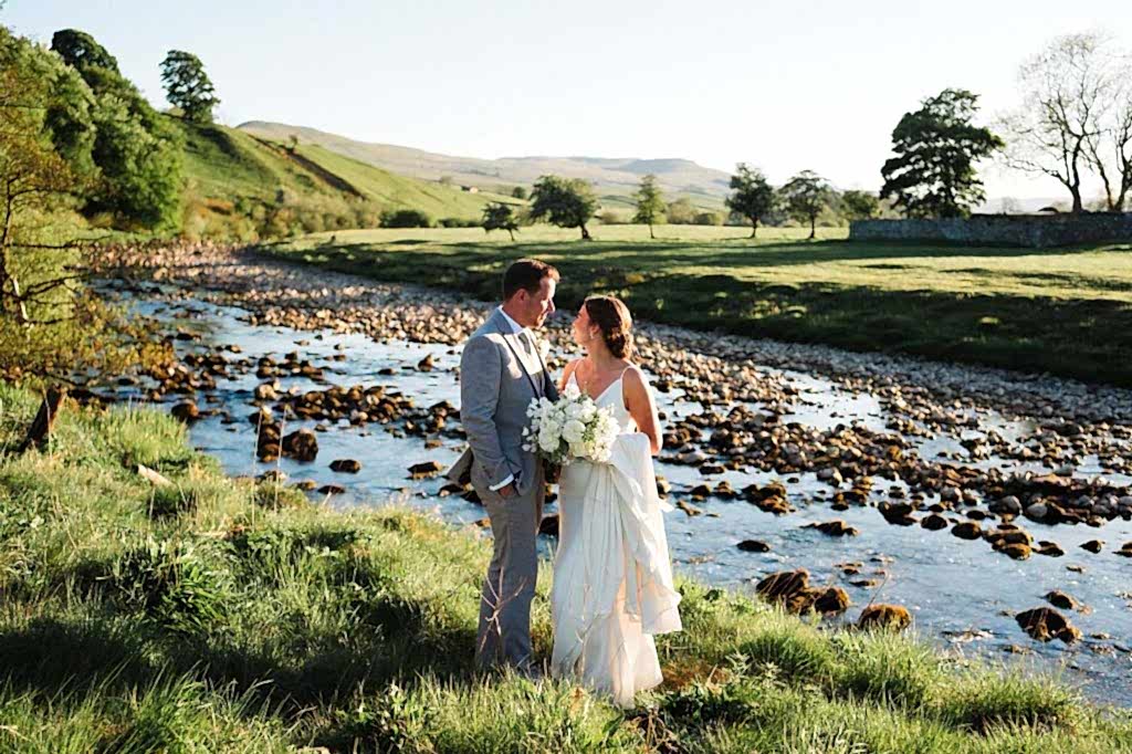 The image shows Melissa and Richard (the bride and groom) posing infront of the Ure River in their wedding attire following their countryside wedding at Yorebridge House.