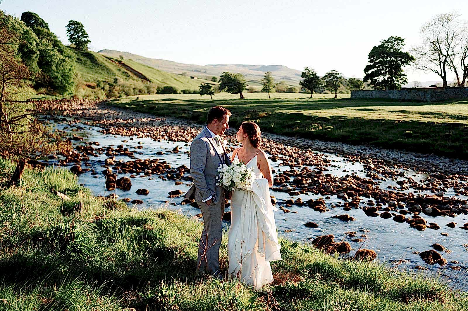 The image shows Melissa and Richard (the bride and groom) posing infront of the Ure River in their wedding attire following their countryside wedding at Yorebridge House.