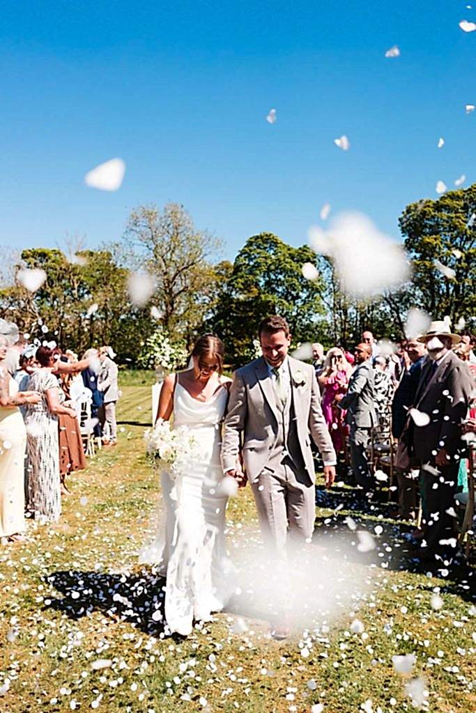 The image shows Melissa and Richard (the bride and groom) walking down the aisle during their exclusive use wedding ceremony at Yorebridge House. The sky is blue, they're surrounded by friends and family and confetti is falling.
