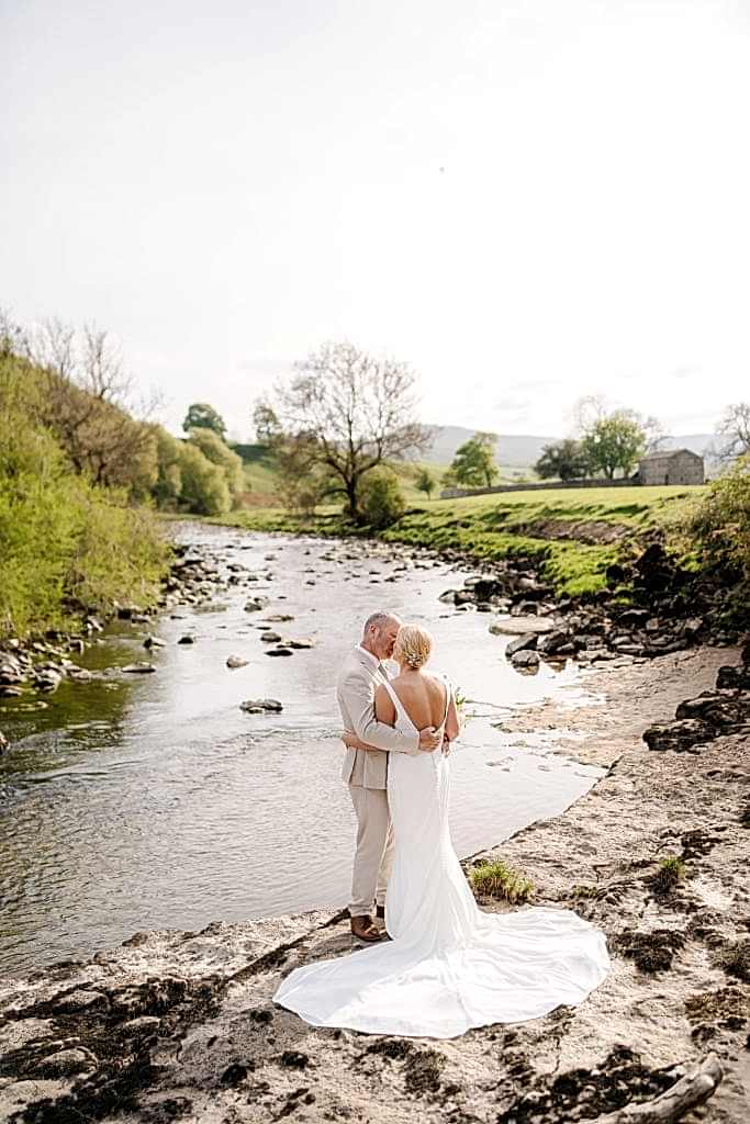 The image shows a bride and groom exchanging a kiss next to the Ure River following their intimate wedding at Yorebridge House. The bride is wearing a fit and flare wedding dress with a low back detailing. Her hair is styled in a low bun with a pearl and diamond hair accessory.
