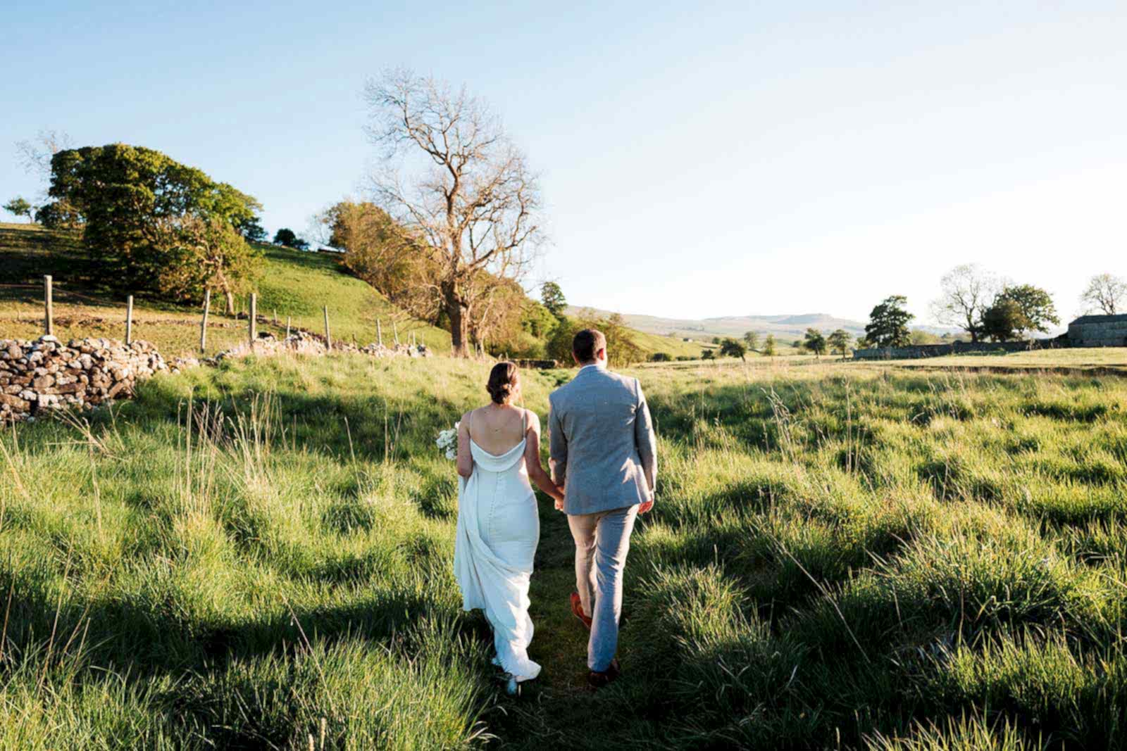 The image shows a bride and groom walking through the countryside at Yorebridge House, holding hands following their Exclusive Use House Wedding in North Yorkshire.