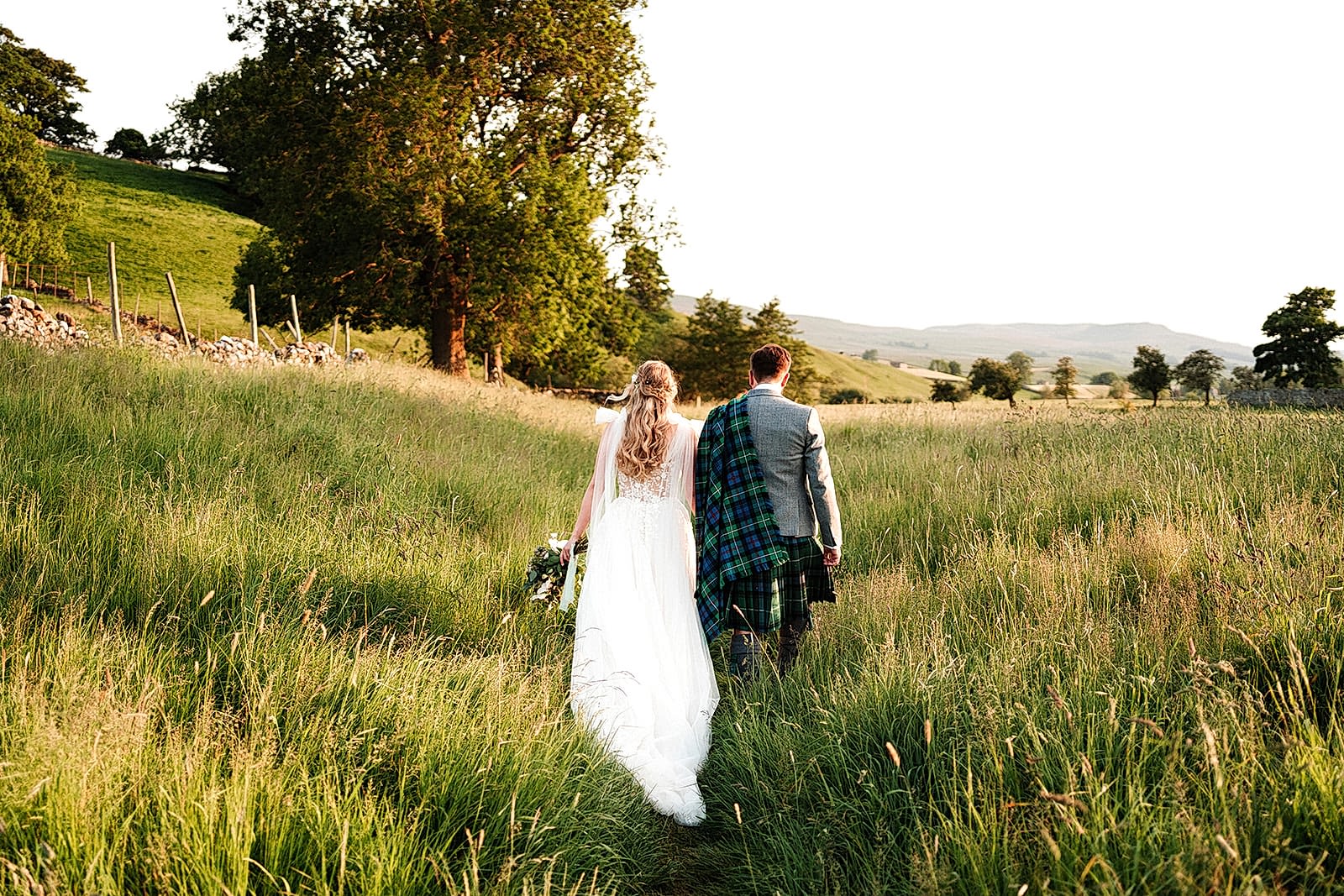 The image shows a bride and groom wandering through the meadows at Yorebridge House in North Yorkshire. Their exclusive use marquee wedding in Bainbridge made for a beautiful wedding celebration.